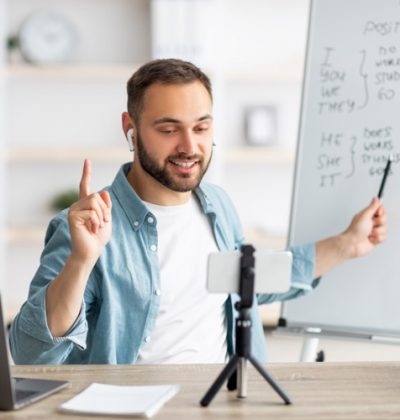 Young male teacher giving online English lesson, pointing at blackboard, explaining grammar rules at home office. Millennial tutor having web conference with pupils, using smartphone for webinar